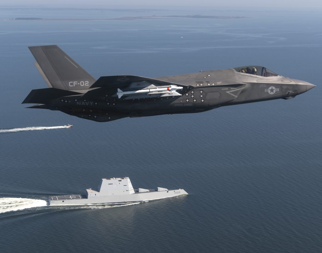 A fighter jet carrier missiles flies over a navy destroyer