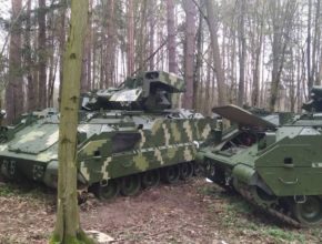 two armored vehicles in camouflage paint sit in the woods