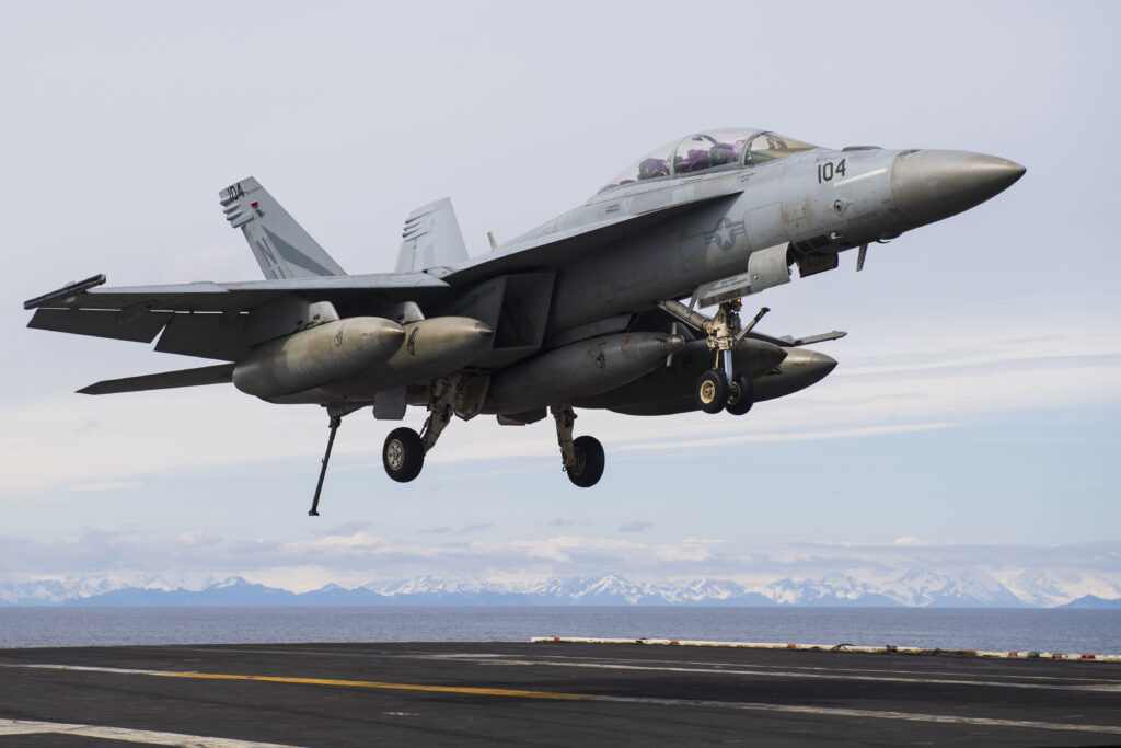 A Navy fighter jet lands on an aircraft carrier with mountains in the background