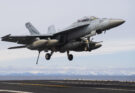 A Navy fighter jet lands on an aircraft carrier with mountains in the background