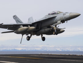 A Navy fighter jet lands on an aircraft carrier with mountains in the background