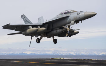 A Navy fighter jet lands on an aircraft carrier with mountains in the background