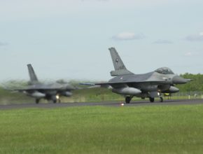 Two fighter jets line up on a runway near a grassy field