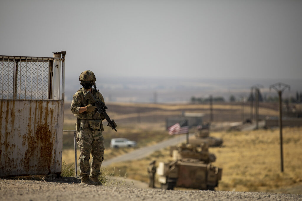 a lone soldier stands at a gate with military vehicles in the background
