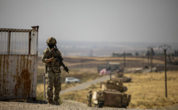 a lone soldier stands at a gate with military vehicles in the background